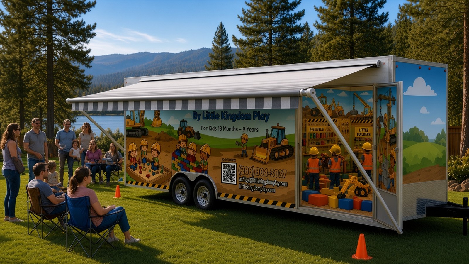 The Little Builders trailer at a community family day on a park lawn by a lake: families relax in the shade of its striped RV-style roll-out awning, traffic cones dot the grass, and kids in hard hats and vests play inside the open side door